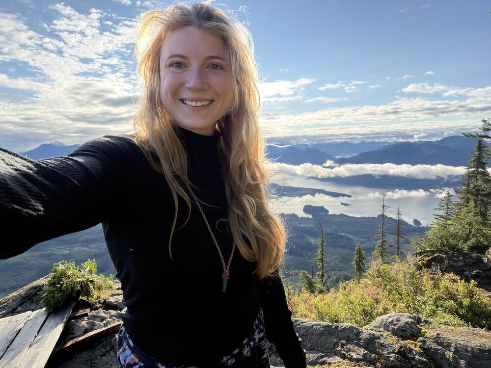 Tessa Cleveland smiling at the top of a mountain peak on the Sunshine Coast, with mountains, an ocean inlet, and a low band of cloud behind her.